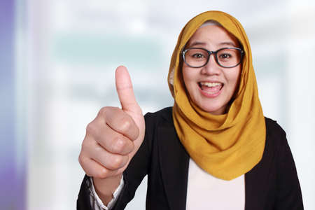 Portrait of beautiful muslim businesswoman wearing hijab showing ok thumb up gesture over blurred defocus background, focus on handの写真素材