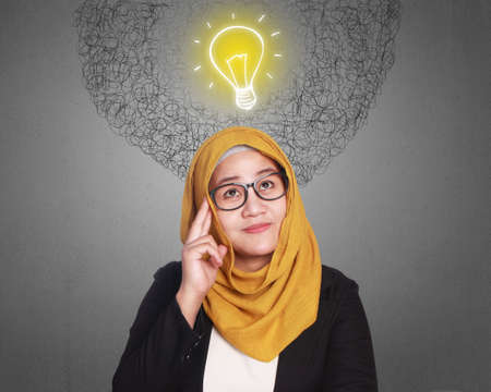 Portrait of muslim businesswoman wearing hijab looking up and smiling thinking idea, bright bulb lamp symbol, problem and solution concept の写真素材