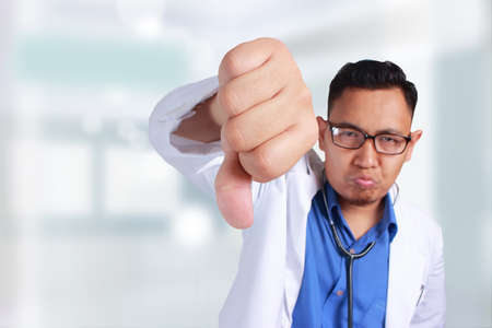 A Young Asian Portrait of a handsome doctor wearing his uniform, showing his thumbs down looking so disappointedの写真素材
