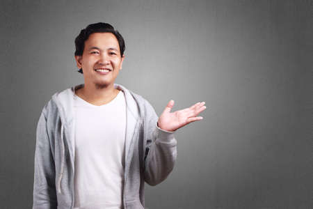 Young Asian man wearing white shirt and gray jacket showing something in his empty hand, copy space. Background on gray. Close up body portraitの写真素材