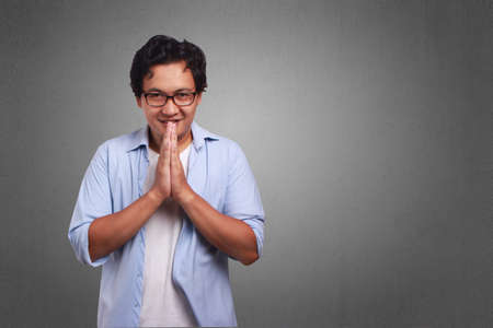 Young Asian man wearing white and blue shirt apologize expression showing clasped hand. Close up...