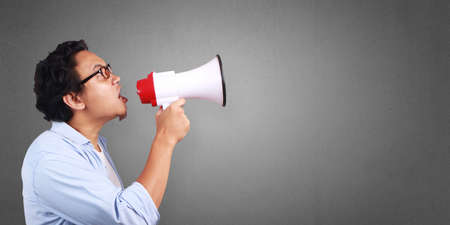 Young Asian man wearing white and blue shirt shouting using megaphone, angry expression. Close up...