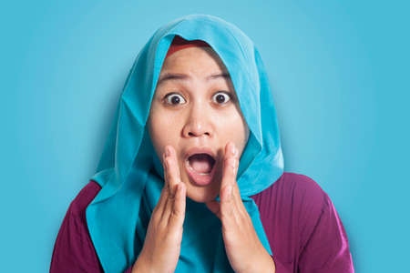 Portrait of beautiful Asian muslim woman wearing hijab shocked with mouth opened over blue backgroundの写真素材