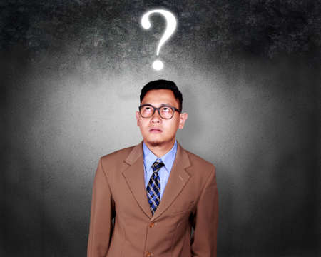Portrait of young confused Asian businessman with question mark, thinking gesture over grunge backgroundの写真素材