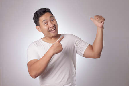 Young Asian man wearing white shirt smiling, showing and pointing something on his side, against white wall with copy space. Close up body portraitの写真素材