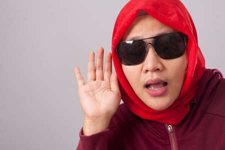 Portrait of Asianmuslim lady in red suit and hijab hold her hand on ear trying to listening carefully to the conversation of someone, curious facial expressionの写真素材