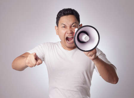 Photo image portrait of funny young Asian man shouting with megaphone, mad yelling screaming crazy supporting motivating conceptの写真素材