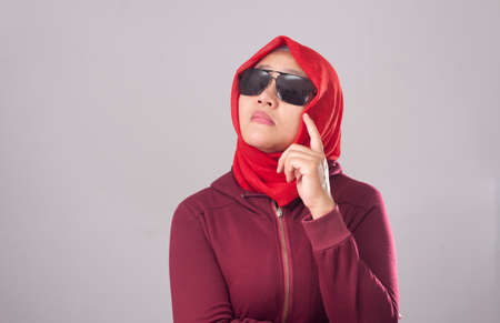 Portrait of young Asian muslim lady wearing black sunglasses in red suit and hijab shows curiousity thinking expression. Looking for idea solution of problemの写真素材