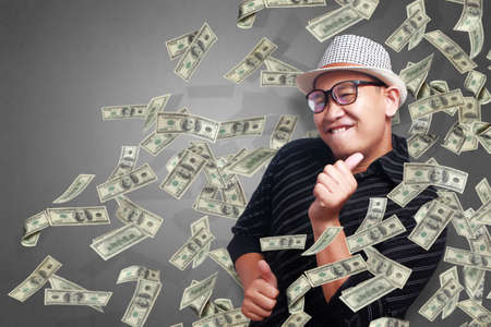 Portrait of happy successful young Asian billionaire man dancing and smiling happily under rain of...