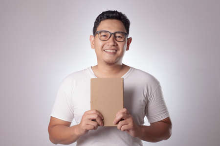 Young Asian man wearing casual white shirt holding a book with blank brown cover, smiling...