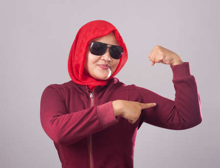 Portrait of Asian muslim lady wearing black sunglasses in red suit and hijab shows strong muscle power gestureの写真素材