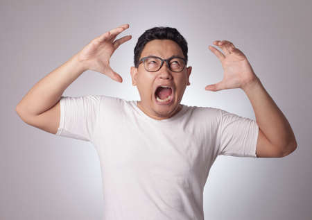 Young Asian man wearing white shirt shouting because of anger. Background on gray. Close up head and shouldersの写真素材