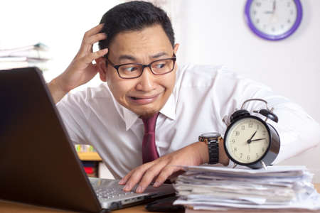 Young Asian businessman worried by time when looking at a clock. Work deadline stress at the officeの写真素材