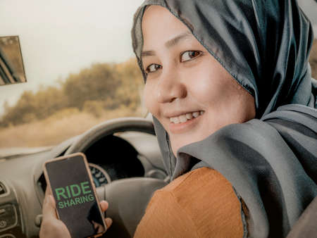 Portrait of Asian muslim woman car ride sharing driver inside her car holding smart phone, looking...