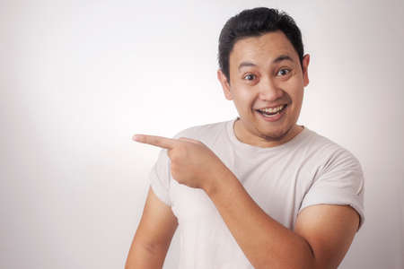 Young Asian man wearing white shirt smiling, showing and pointing something on his side, against...