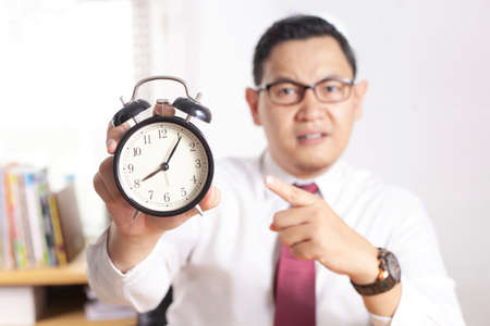 Young Asian businessman pointing at clock, angry expression. Time deadline concept. Man working in...