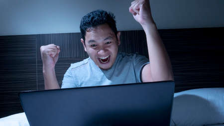 Young Asian man wearing casual shirt looking at laptop on bed at the midnight, winning gesture....