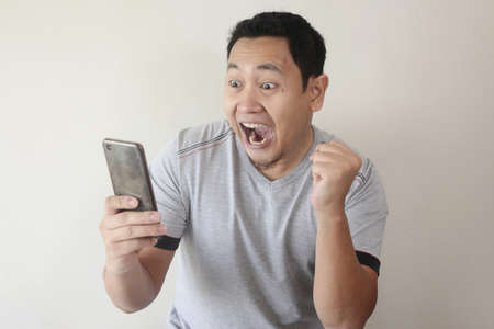 Portrait of happy funny Asian man shocked or surprised with mouth open, good news on smart phone concept, amazed winning gestureの写真素材