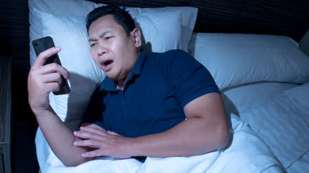 Receiving bad news on phone at midnight concept, Asian male shocked annoyed expression to have bad news on phone while lying on bed at midnightの写真素材