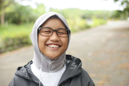 Portrait of happy Asian muslim teenager girl using earphone listening to music on her smart phone in the parkの写真素材