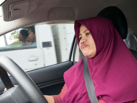 Portrait of funny Asian female muslim driver get bored in her car trapped in traffic jam, tired lazy...