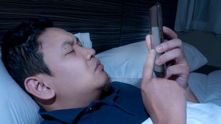 Phone addicted concept, young Asian man using smart phone on bed at midnight, reading chat message,...