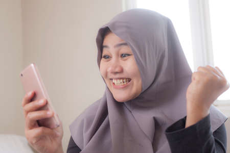 Portrait of young Asian muslim businesswoman get good news on her phone, happy surprised expressionの写真素材