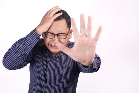 Portrait of crying sad young Asian man showing surrender gesture, hands raised up asking to stop...