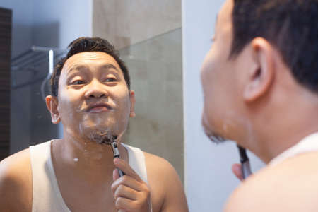 Portrait of attractive young Asian man shaving in bathroom, smiling and looking at mirror