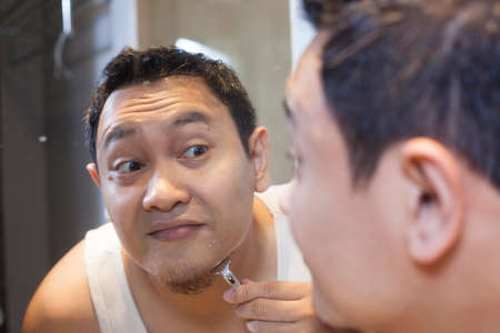 Portrait of attractive young Asian man shaving in bathroom, smiling and looking at mirror