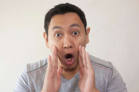 Portrait of Young asian men in casual grey shirt, shocked gesture with open mouth. Hands rised up