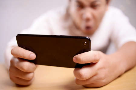 Photo image portrait of a cute handsome young Asian man with funny face playing games on tablet, sad...