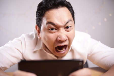Photo image portrait of a cute handsome young Asian man with funny face playing games on tablet, sad angry loosing expressionの写真素材