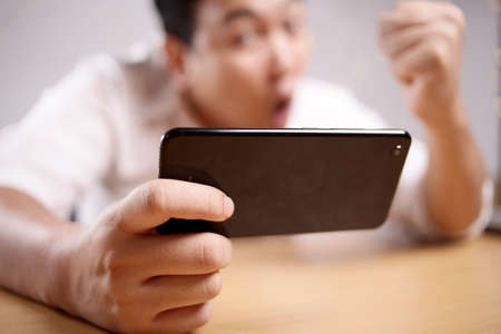 Photo image portrait of a cute handsome young Asian man with funny face playing games on tablet...