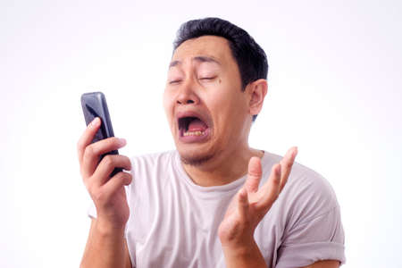 Young Asian man crying shocked worried expression by the phone. Over white backgroundの写真素材
