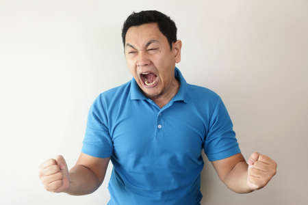 Young Asian man wearing blue shirt shouting because of anger. Against grey wall. Close up head and shouldersの写真素材