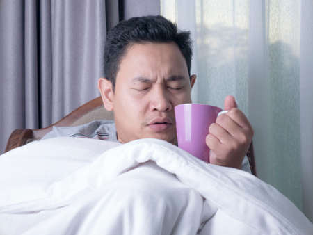 Sick cold young Asian man covered with blanket while sitting on chair, drinking hot drinkの写真素材
