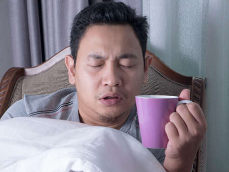 Sick cold young Asian man covered with blanket while sitting on chair, drinking hot drinkの写真素材