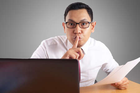 Portrait of young Asian businessman asked to be silenced, not to tell any secret, shushing gesture, closing lips mouth with fingerの写真素材