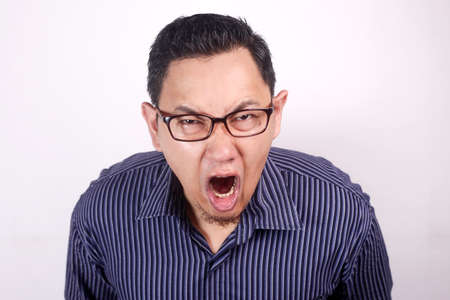 Young Asian man wearing blue shirt shouting because of anger. Against white wallの写真素材