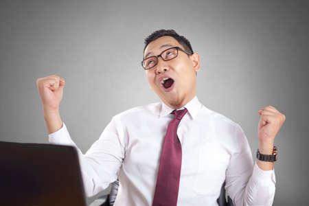 Portrait of young attractive Asian businessman working on laptop at the office, smiling happy expression winning gesture, celebrating victoryの写真素材
