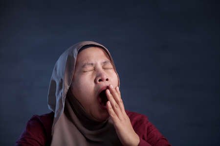 Young Asian muslim woman wearing hijab yawning tired, dizzy expression. Close up head and shoulders over dark blue backgroundの写真素材