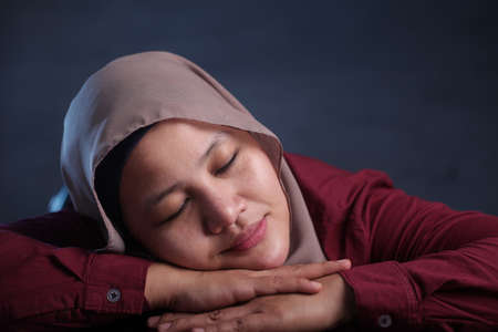 Young Asian businesswoman wearing hijab tired and falling asleep on desk at work, smiling and dreaming expression. Close up head and shoulders against dark backgroundの写真素材