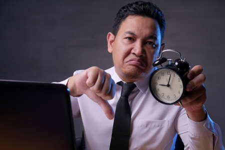 Young Asian businessman showing a clock and thumbs down, angry expression. Time deadline concept. Man working in officeの写真素材