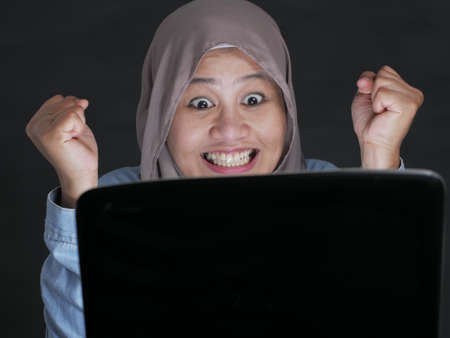 Portrait of Asian muslim lady using laptop, shows happy surprised expression celebrating winning victory gesture after receiving good news on her emailの写真素材