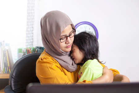 Asian muslim single mother calms her little baby daughter at work, freelancer woman working from homeの写真素材