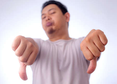Photo image portrait of a funny young Asian man doing mocking gesture with synical face and showing two thumbs down over white backgroundの写真素材