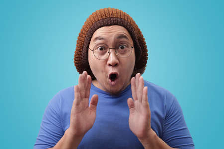 Portrait of young funny Asian man shocked or surprised expression with mouth open, against blue backgroundの写真素材