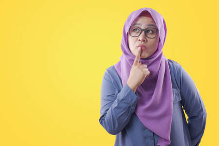 Asian muslim woman wearing hijab with thinking expression. Looking for solution of a problem. Close up portrait against yellow backgroundの写真素材