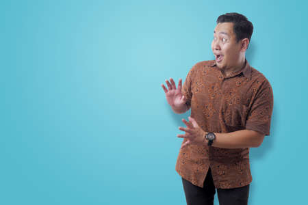 Portrait of young Asian man wearing Indonesian batik shirt shocked to see something on his side,...